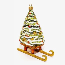 A hand-painted glass Christmas ornament shaped like a snow-covered tree on a sleigh, featuring green and white layers with gold, red, and yellow ornaments, set against a plain white background.