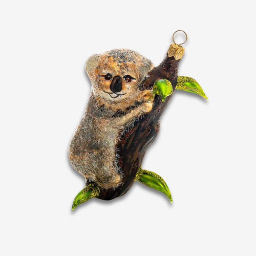 A handcrafted glass koala ornament clinging to a tree branch with green leaves, featuring a gold hanging loop at the top, set against a plain light gray background.