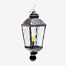 A hanging lantern ornament with a black metal frame, textured clear glass panels, a small yellow light inside, and a silver hanging loop on a plain light gray background.