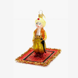 A hand-painted glass Christmas ornament depicting a man in traditional Middle Eastern attire standing on a red and gold patterned rug, set against a plain white background.