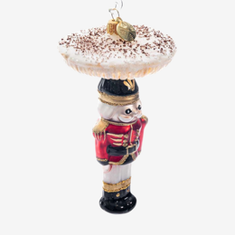 A nutcracker-shaped ornament carrying a large frosted beverage on its head, decorated with a red uniform, gold trim, and a gold hanging loop at the top, shown on a plain light gray background.