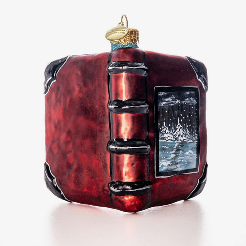 A red book-shaped Christmas ornament with black spine details, a small window showing a snowy night scene, and a gold hanging loop on a white background.