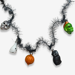 A Halloween garland necklace featuring five classic ornaments: a silver skull, a white ghost, an orange pumpkin, a black cat, and a green Grinch head, all strung on a black tinsel chain against a light gray background.
