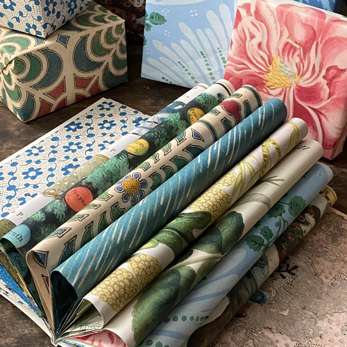 A stack of rolled and folded patterned wrapping paper sheets displayed on a rustic wooden surface, with additional patterned paper pieces from the John Derian Wrapping Paper Book partially visible in the background.