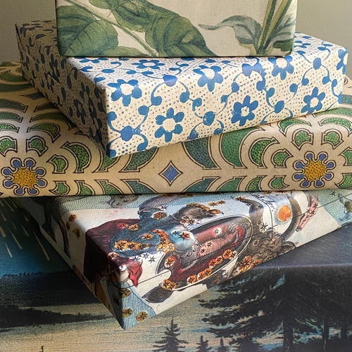 Three stacked rectangular boxes wrapped in decorative printed paper. The top box has green tropical leaves on a cream background, the middle box has blue flowers with white dots on a cream background, and the bottom box has a vintage car illustration with trees and sky.