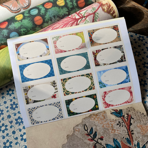 A printed sheet of oval label stickers with blank white centers, placed over a patterned fabric surface. The sheet contains multiple small rectangular patterned label designs, and other patterned fabric rolls are visible around it.