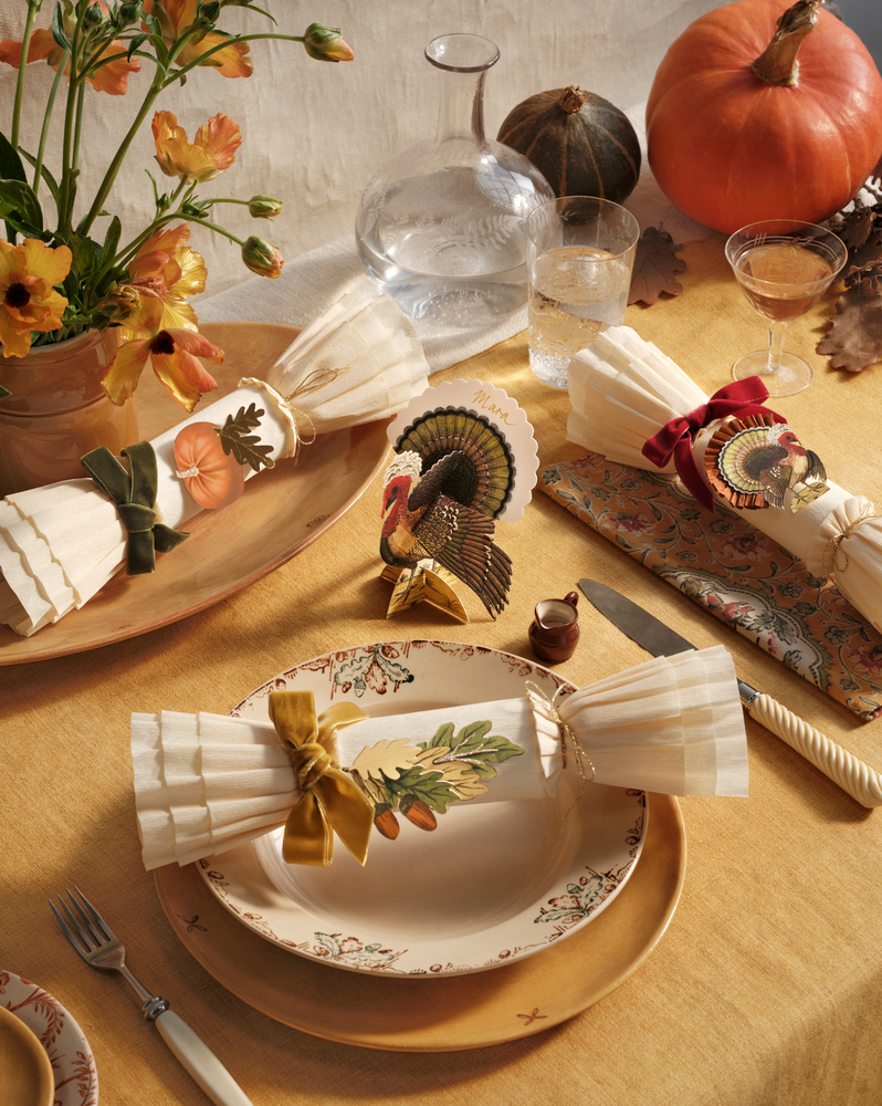 An autumn-themed table setting with a beige tablecloth, patterned plates, napkins tied with bows, a decorative turkey figurine, pumpkins, and floral accents.