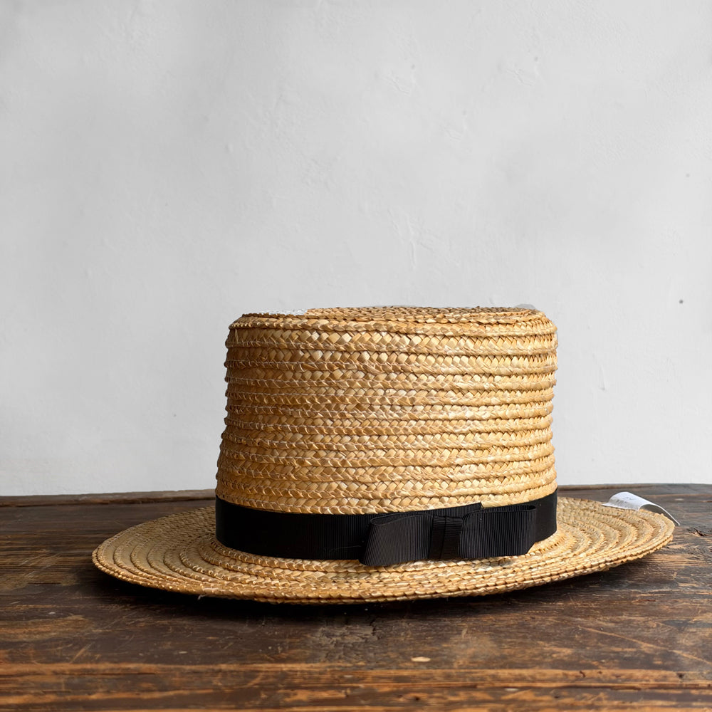 A natural straw amish hat with a black ribbon band and bow, resting on a dark wooden surface against a light gray wall.