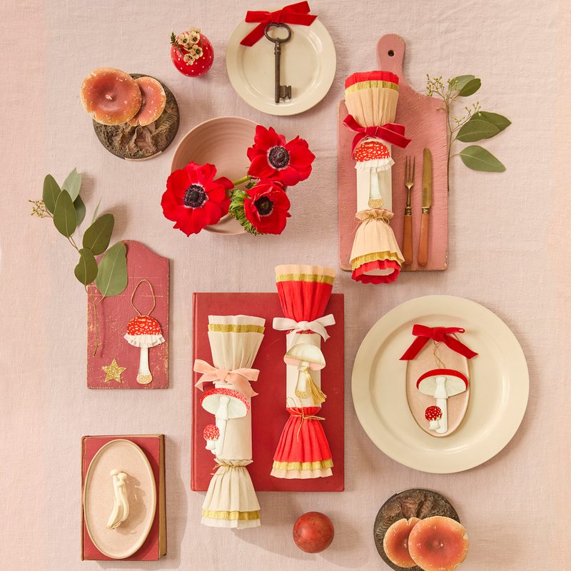 A styled flat-lay of autumn-themed tableware and decor on a pale blush background, including red-and-cream ceramic plates, mushroom-shaped cutlery, red poppy flowers, small candles, a key, and greenery.