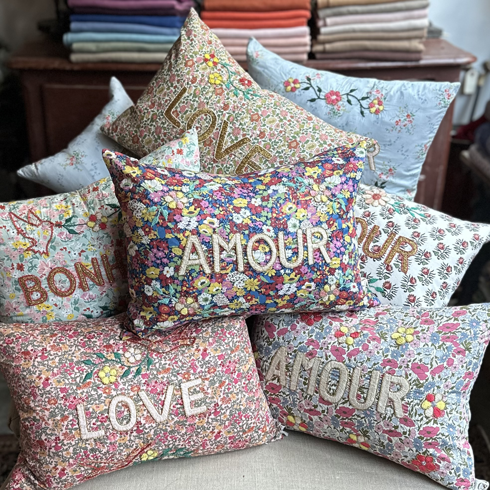 A stack of colorful pillows featuring floral patterns and motifs with hand stitched "Amour" or "Love" embroidered onto the front